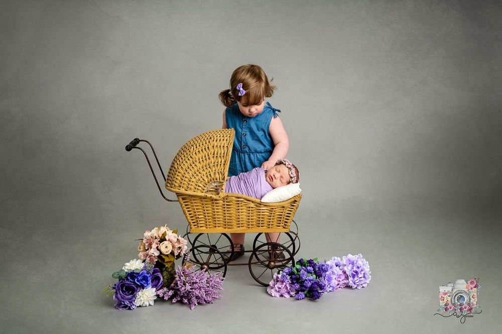 Vintage wicker baby carriage styled photo setup with blue dress and purple flowers against grey background.