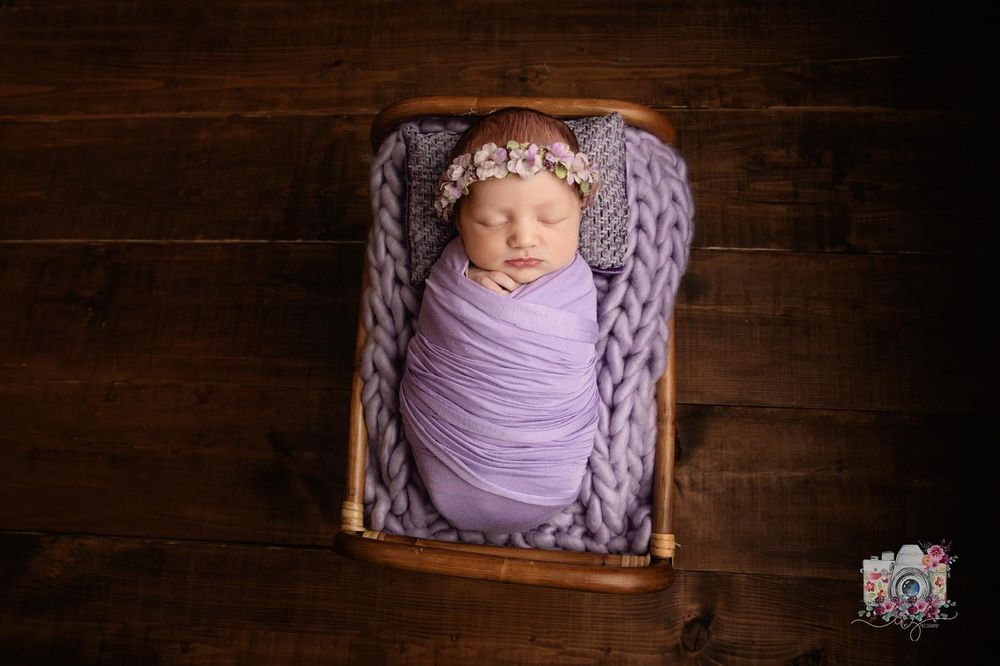 Newborn in lavender wrap and headband nestled in knitted blanket on wooden backdrop.