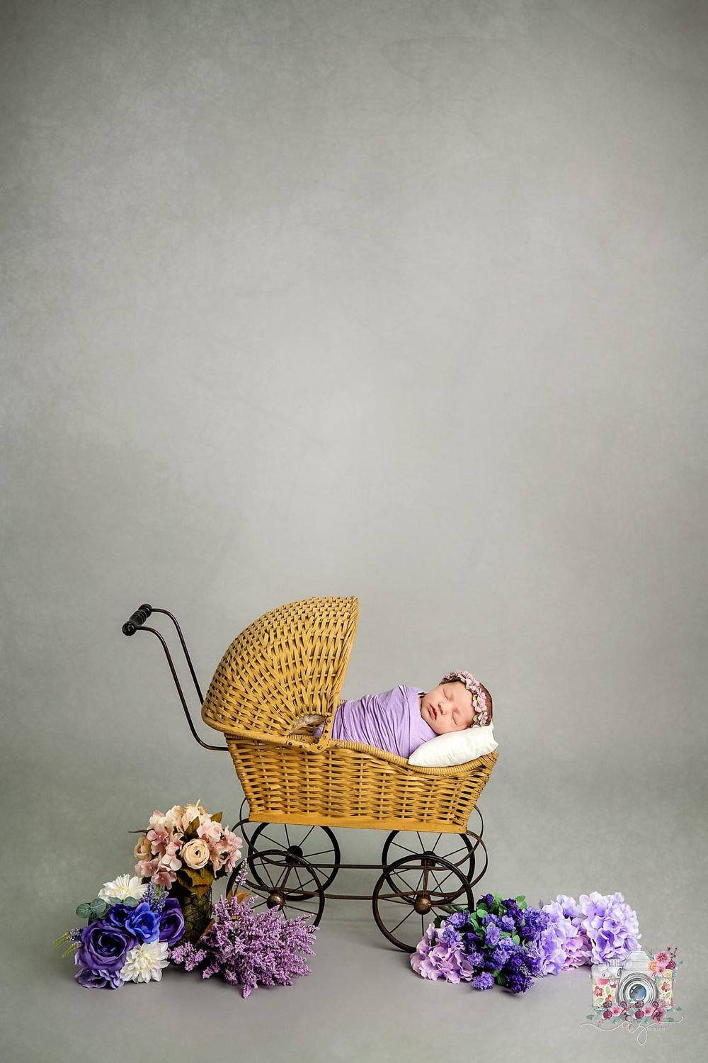 Vintage wicker baby carriage with purple flowers and newborn in lavender wrap against grey backdrop.
