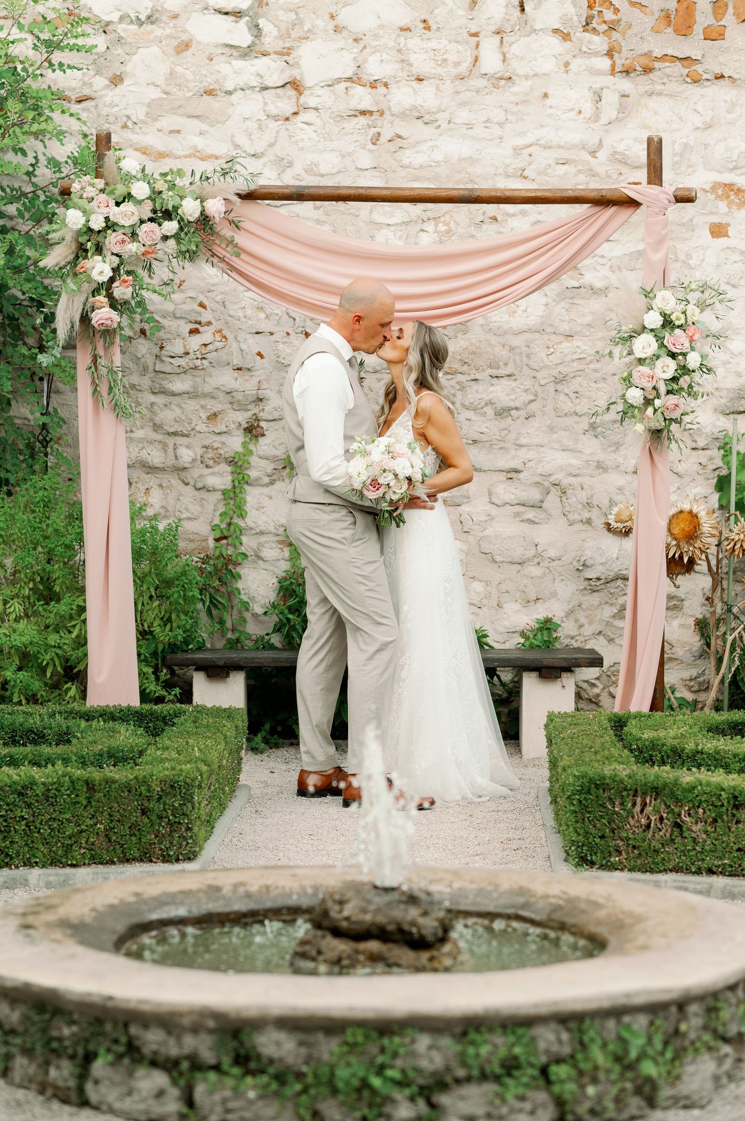 Romantic wedding moment captured under a draped pink arch with stone wall and fountain.