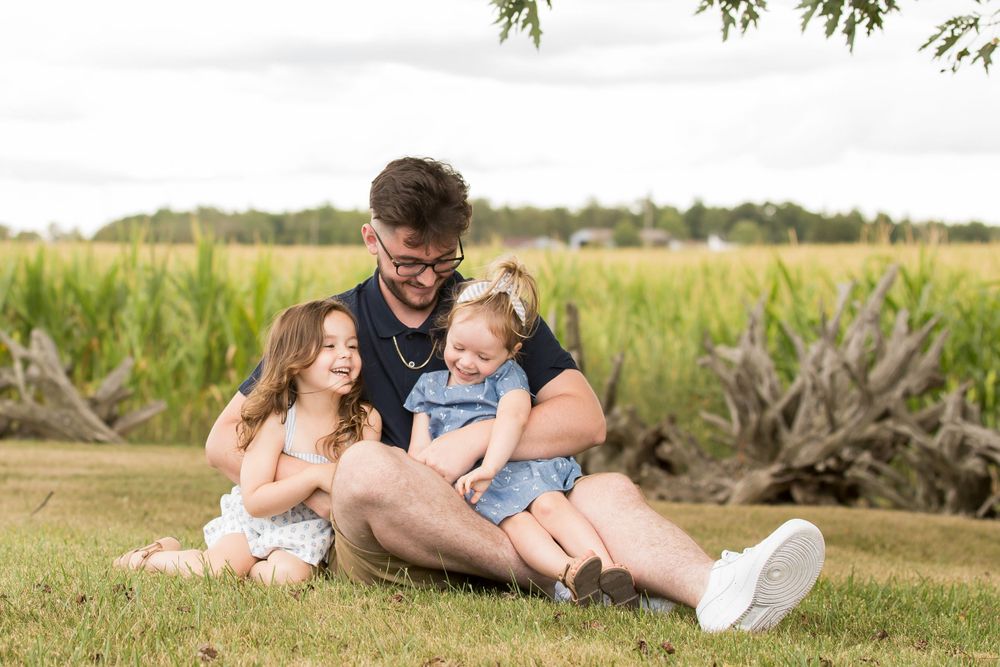 Big brother and little sisters sit together and laugh on grass with lush greenery and trees in the background.