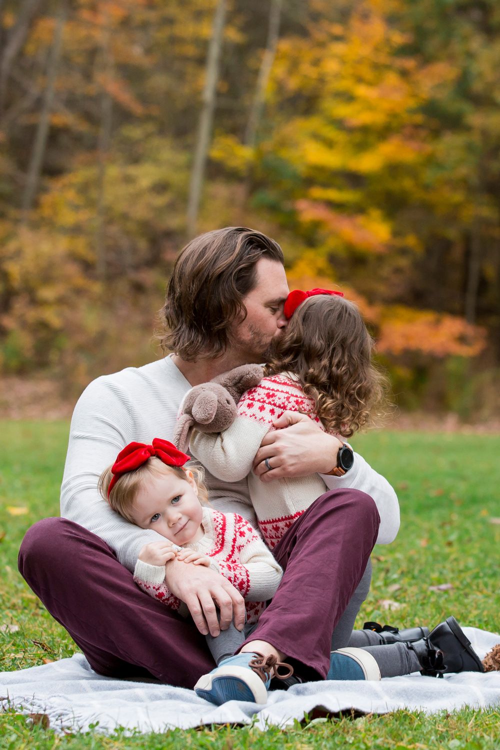 A sweet moment captured during an Fall photoshoot on a blanket with fall foliage in the background of dad hugging and kissing his daughters.