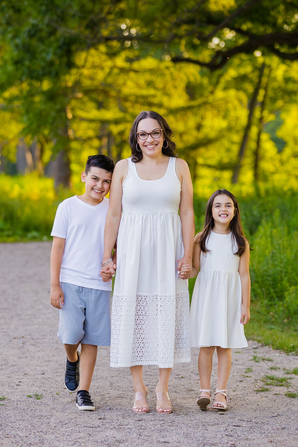 A mother and her two kids wearing white outfits walks together on a dirt path surrounded by lush summer foliage.