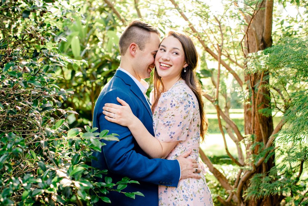 A couple embraces in a garden setting with lush greenery, the person in navy suit with partner wearing a floral dress.
