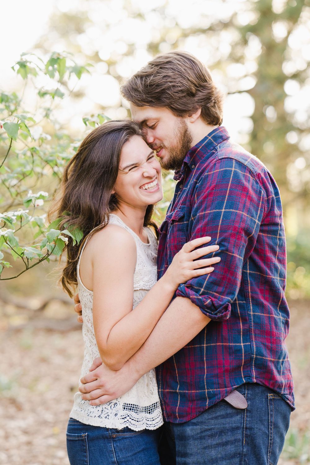 A romantic couple embraces outdoors in casual attire against a soft natural background with green foliage.