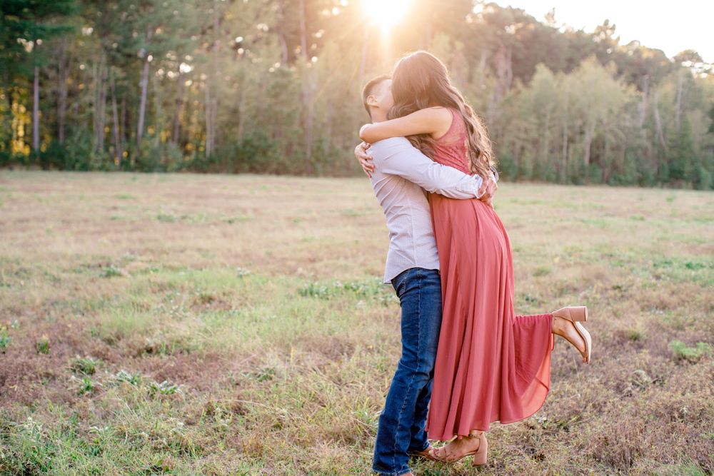 Two people embrace in a sunlit field at golden hour as her coral maxi dress flows in the breeze.