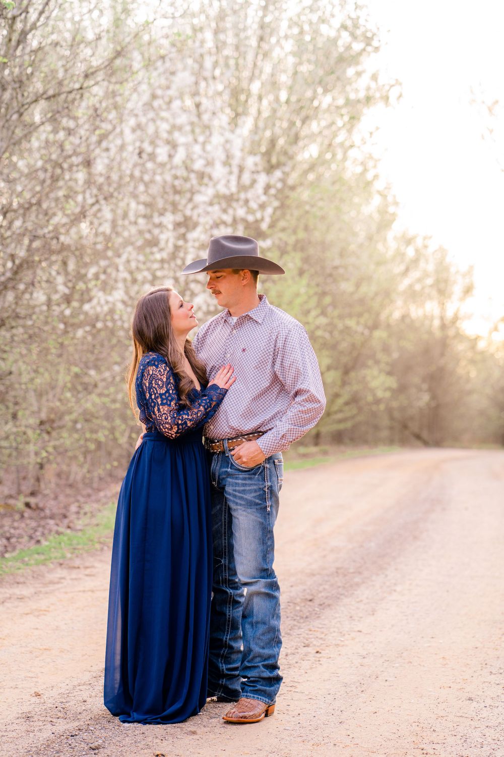 Romantic couple in western attire standing on a sunlit dirt road during golden hour.