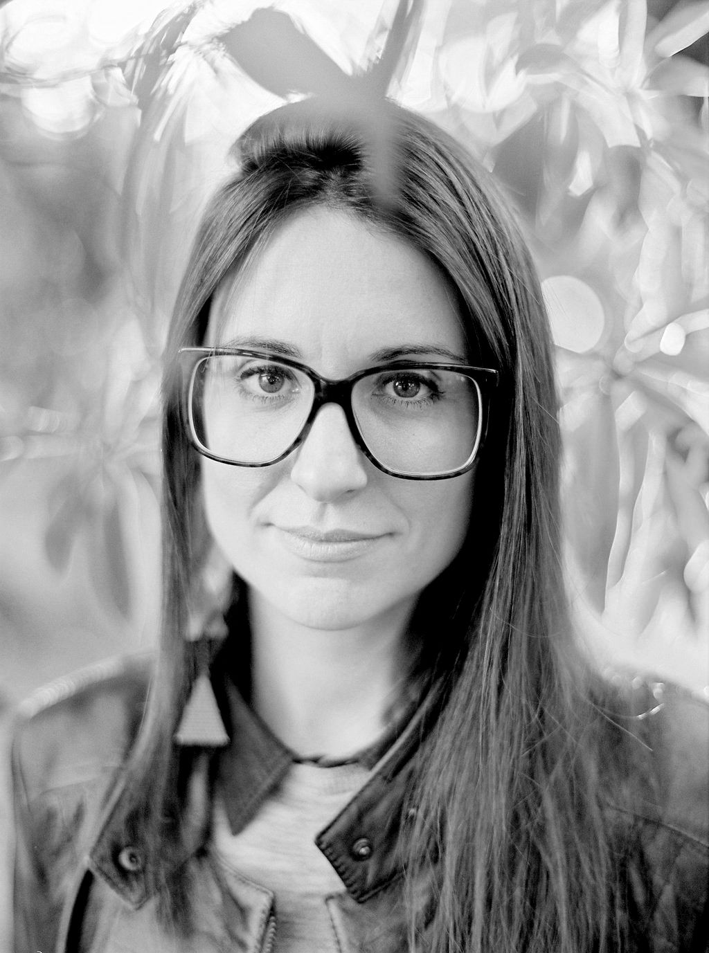 Black and white portrait featuring glasses and long straight hair with a soft smile in a natural setting.