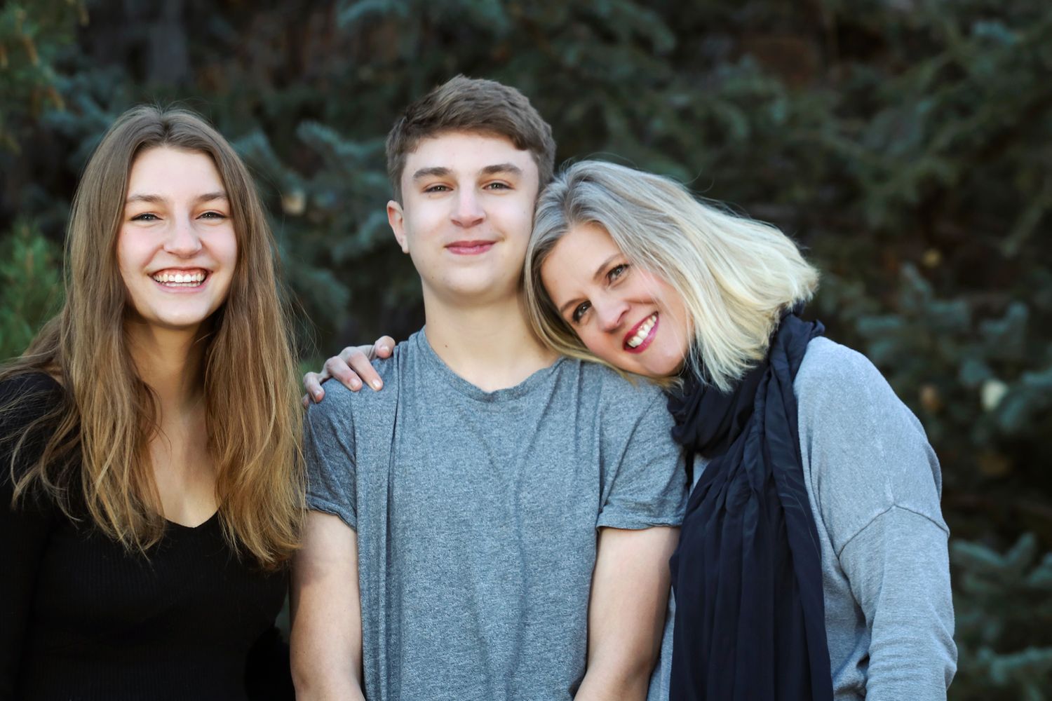 Three people smiling together outdoors with dark foliage in the background.