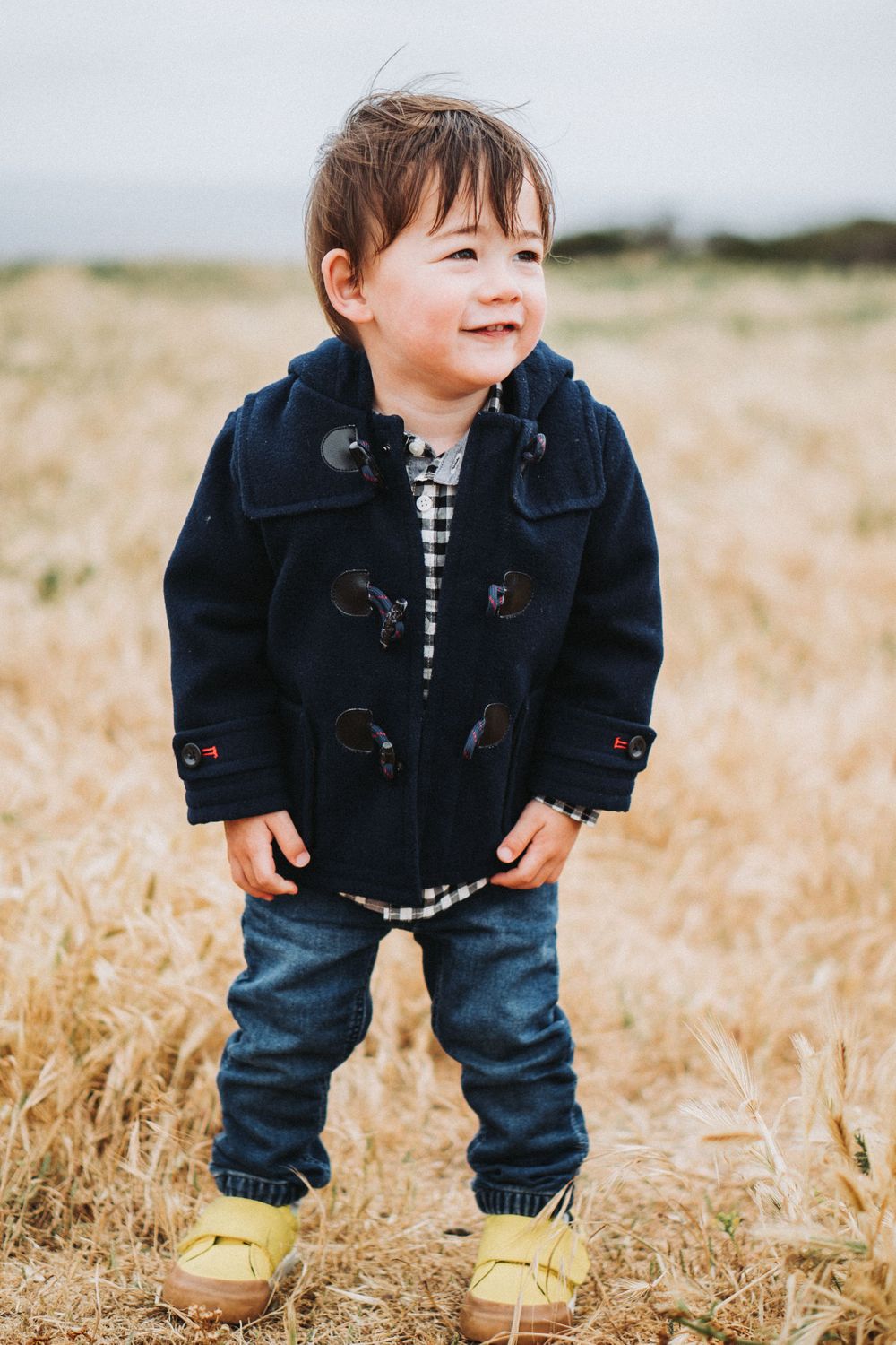 Cute toddler boy in navy duffle coat and yellow sneakers standing in golden wheat field.