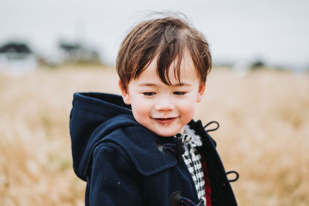 Adorable young boy in navy hooded coat smiling softly while standing in golden wheat field.