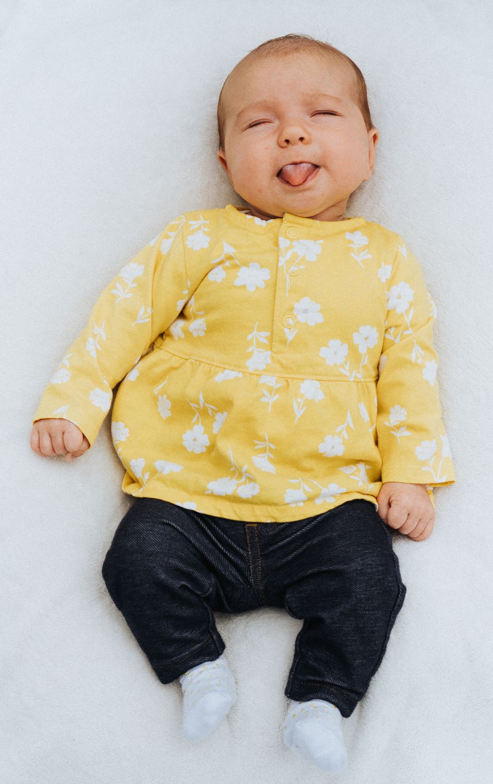 Adorable baby in yellow floral top and dark jeans lying on white surface sticking tongue out playfully.