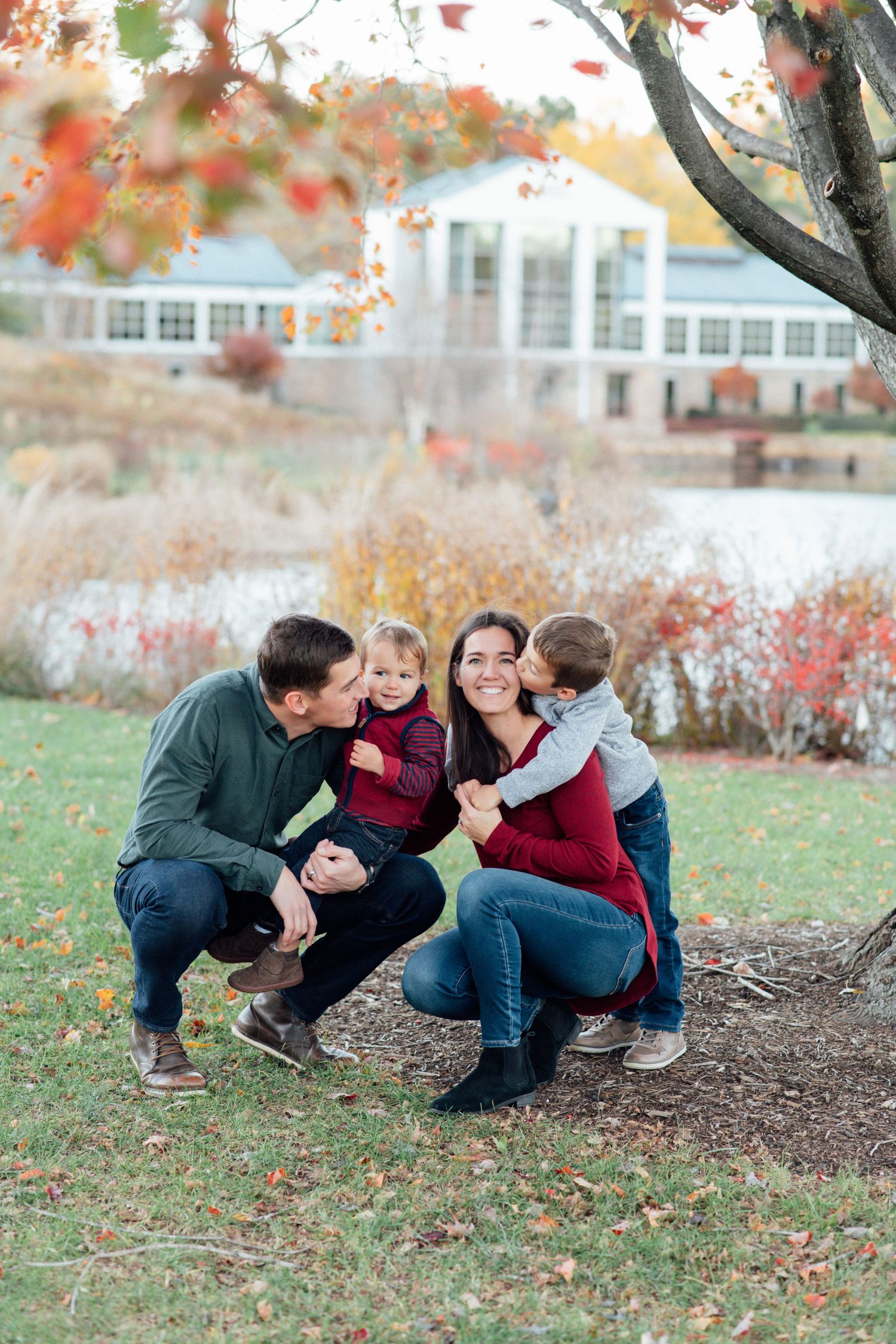 Fall family portrait outdoors with colorful autumn foliage and white building in background.