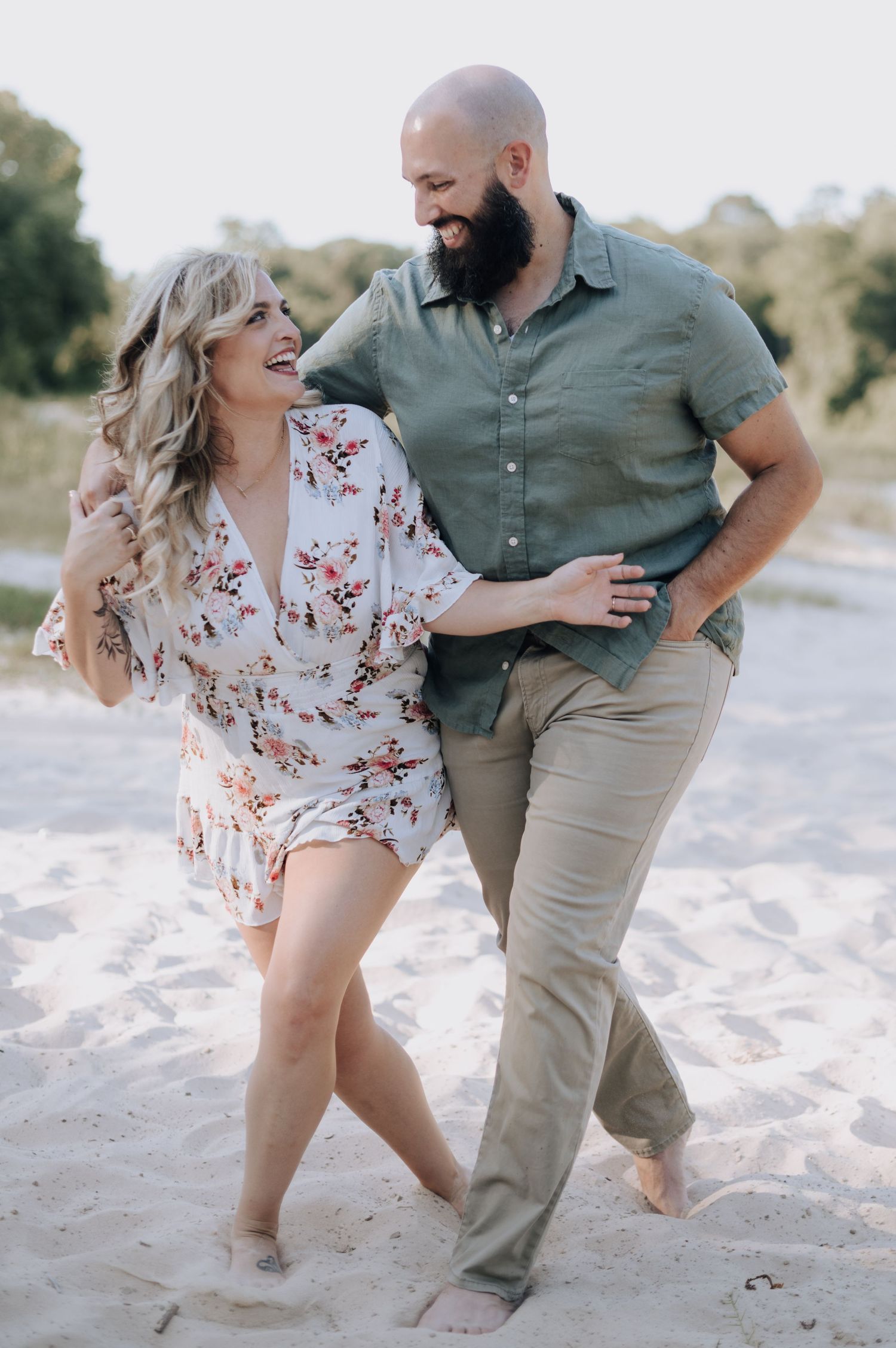 A joyful couple wearing casual attire laughs together while walking barefoot on a sandy beach.