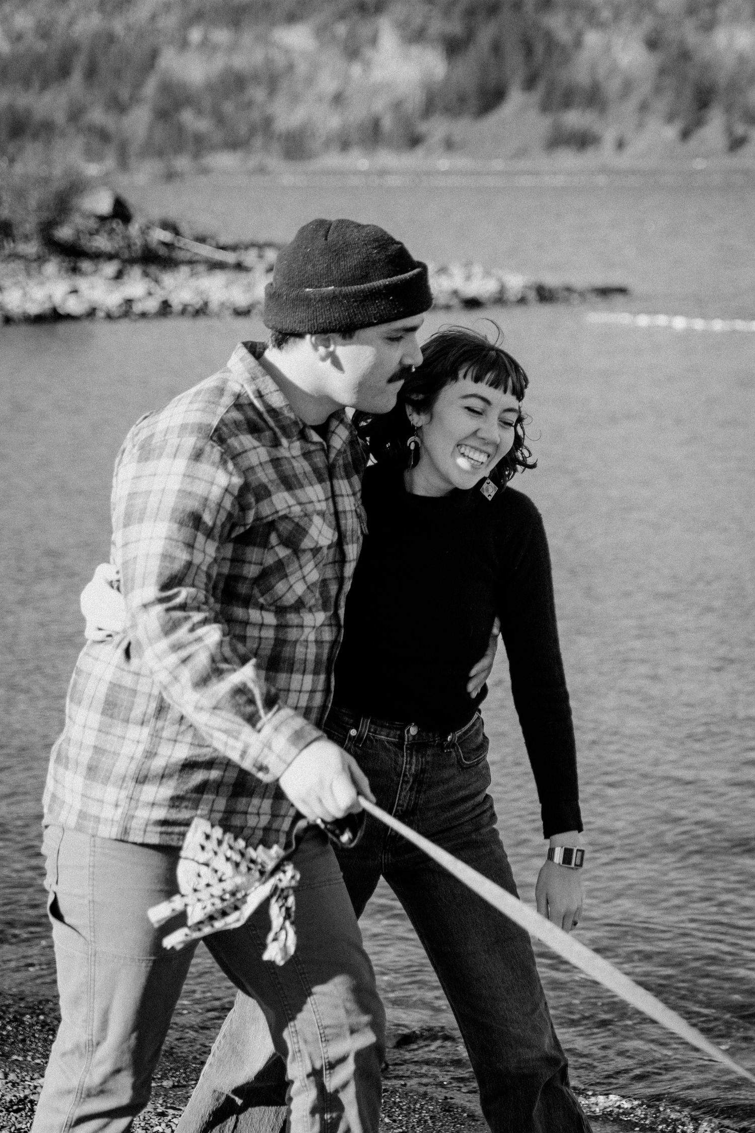 A couple shares a playful moment by the water while one person holds a wooden oar.