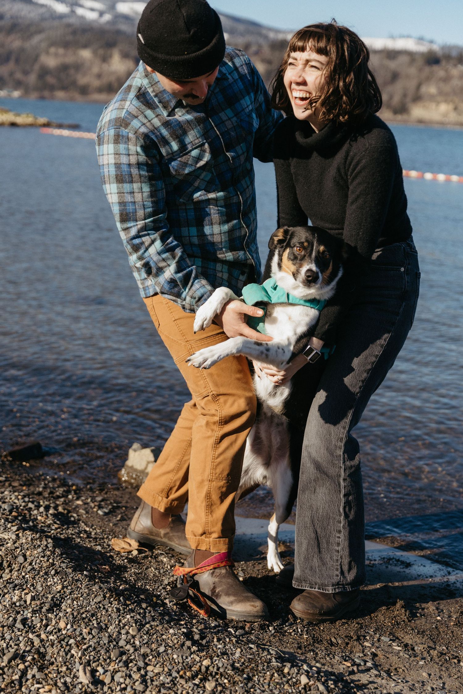 Happy couple playing with their dog by the water's edge on a sunny day.