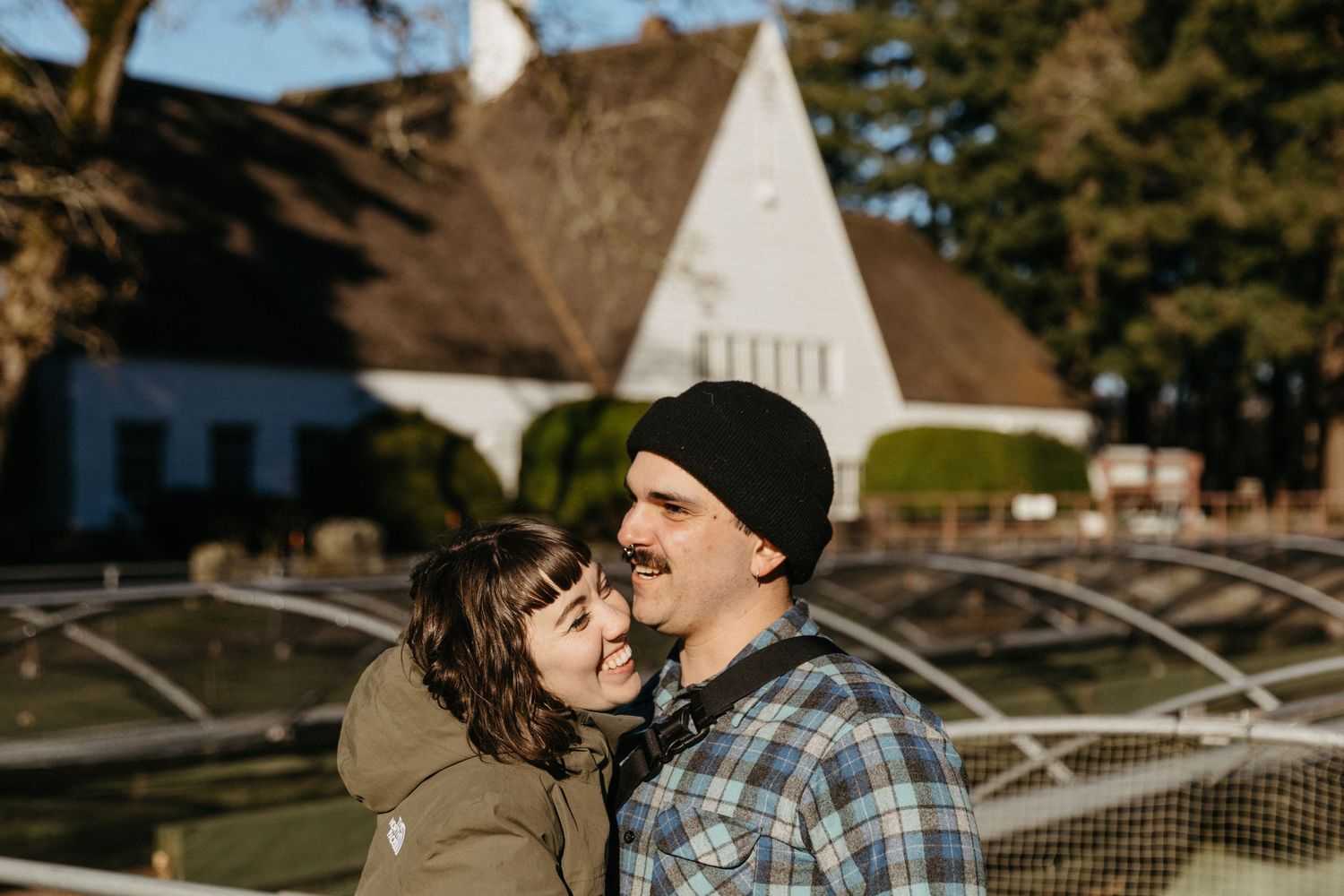 Couple sharing a tender moment near white A-frame buildings and greenhouse structures.