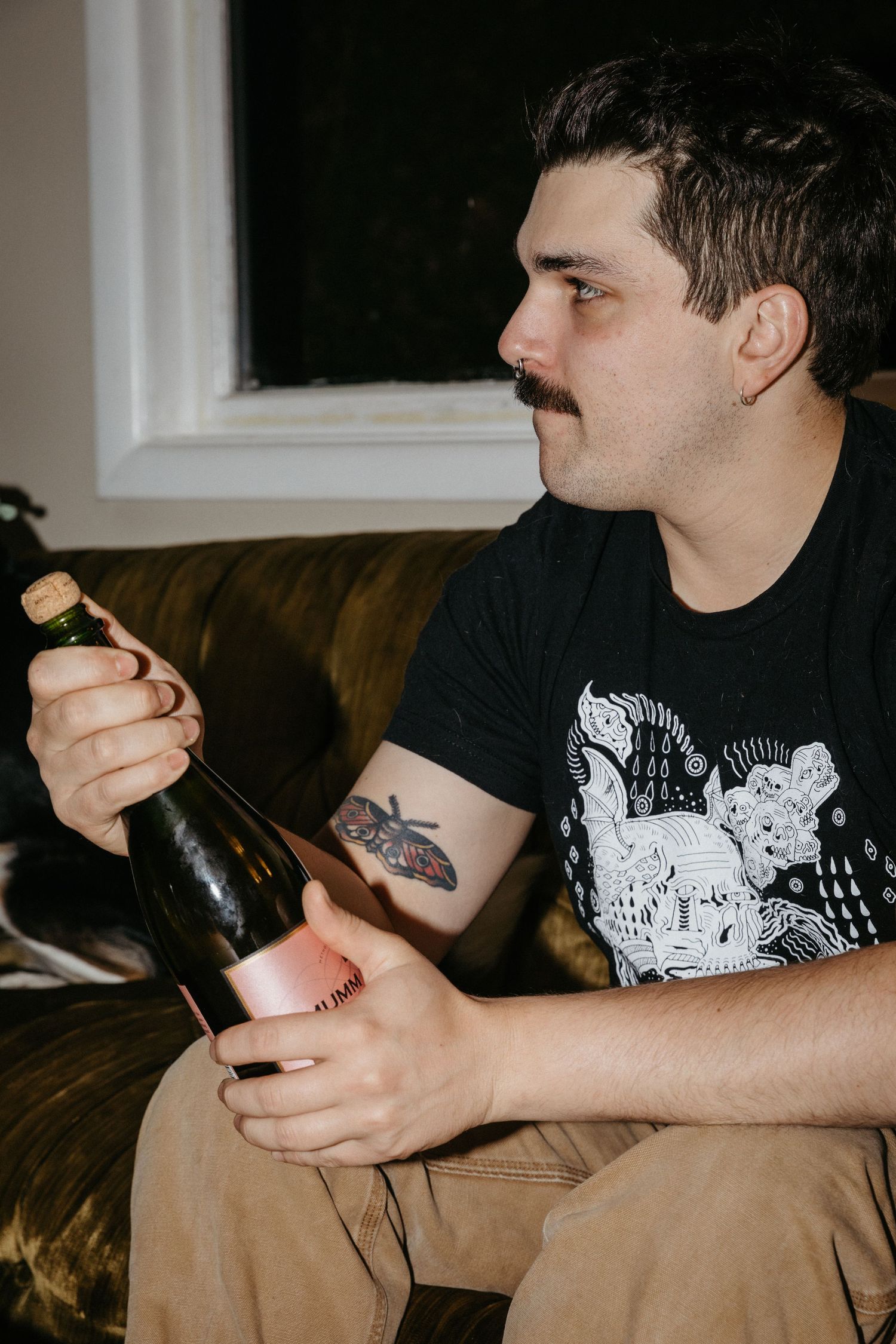 Person with tattoo opening a bottle while sitting on vintage velvet couch.