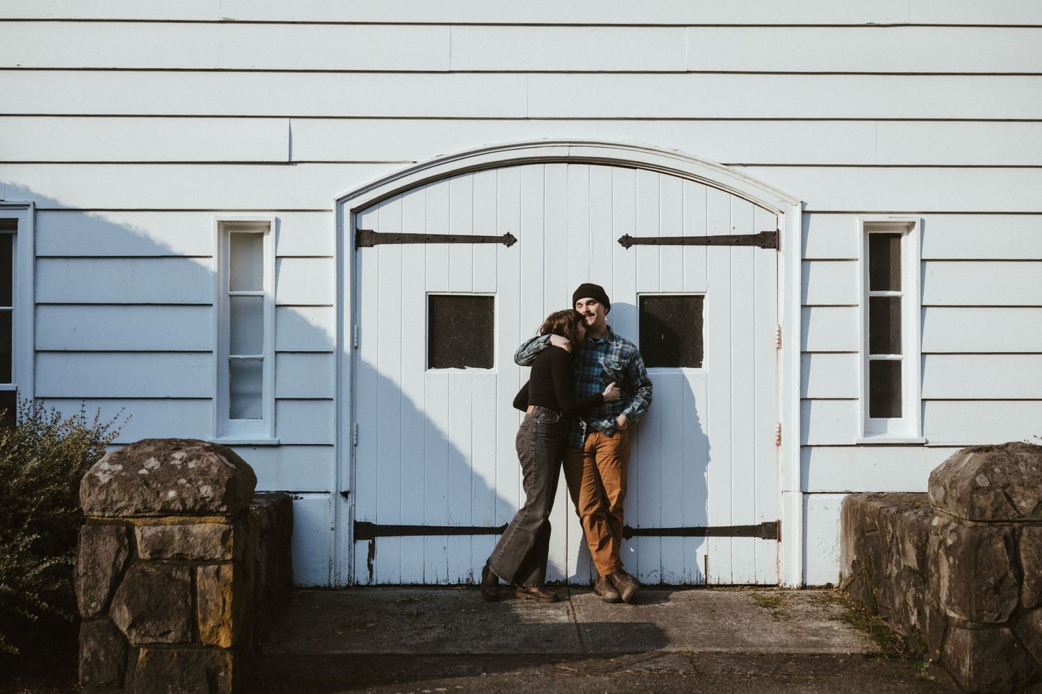 Two people pose against a white barn door with black hinges between stone pillars.