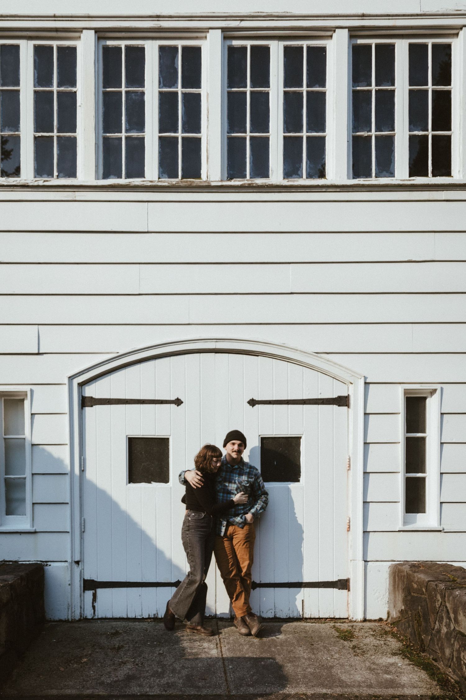 A couple stands against a white barn door with decorative hinges and windows above.