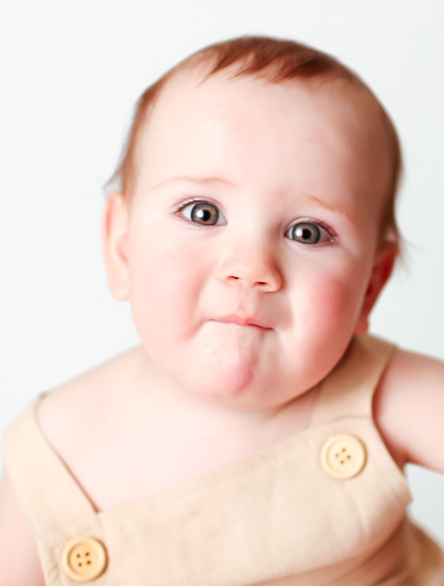A cute baby with wide eyes and chubby cheeks wearing a beige outfit with buttons against a white background.