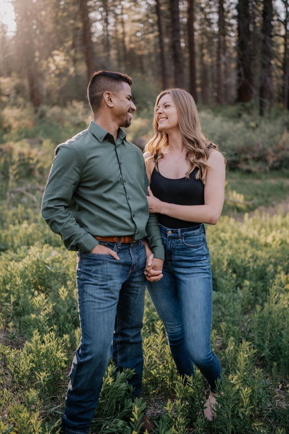 Smiling couple holding hands and gazing at each other in golden sunlit woodland clearing.