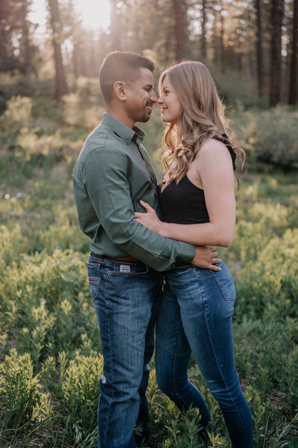 Happy couple embracing and smiling at each other in sunlit forest meadow during engagement shoot.