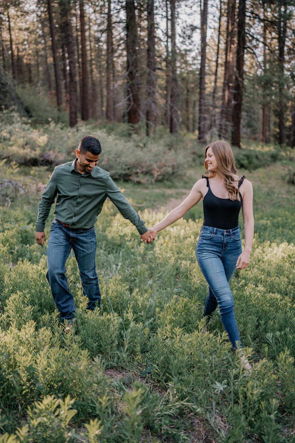Couple walking hand in hand through lush green forest meadow during outdoor engagement session.