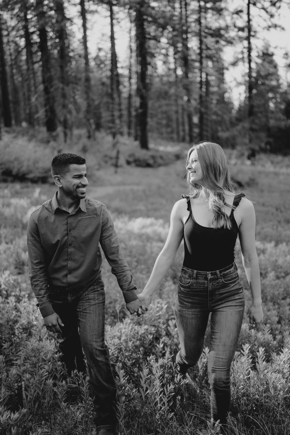 Black and white photo of laughing couple holding hands while walking through forest clearing.