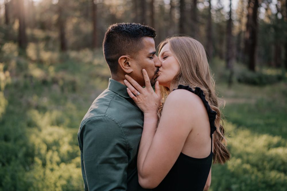 Couple sharing a kiss in sun-drenched forest clearing during romantic outdoor engagement session.