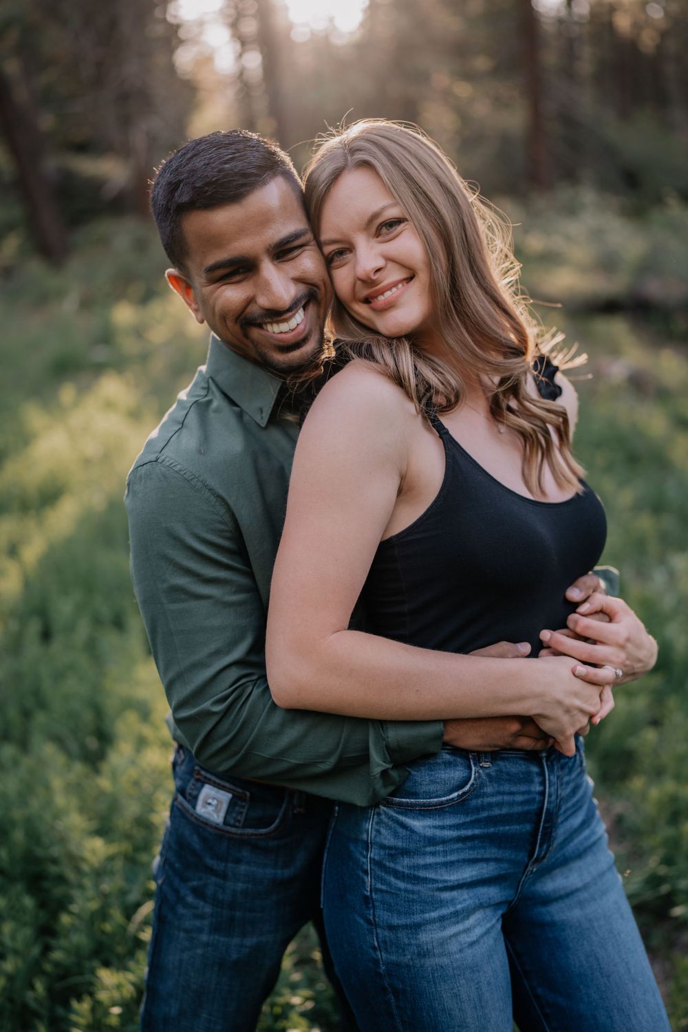 Joyful couple hugging and smiling at camera in sunlit green forest engagement portrait.