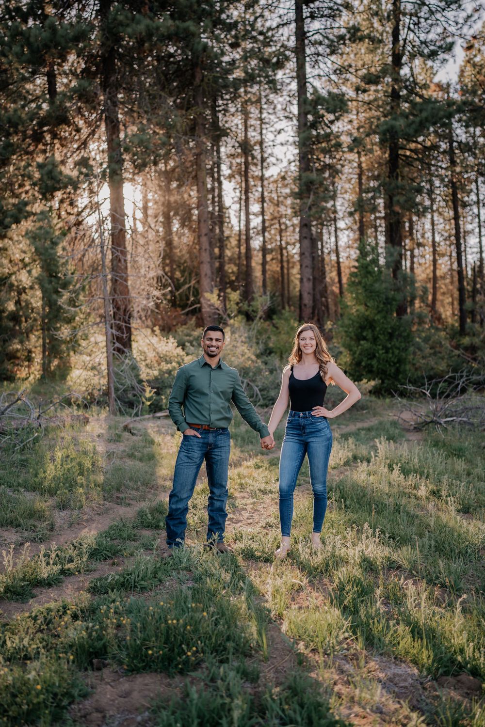Couple holding hands smiling in a sunlit pine forest during golden hour engagement photo session.