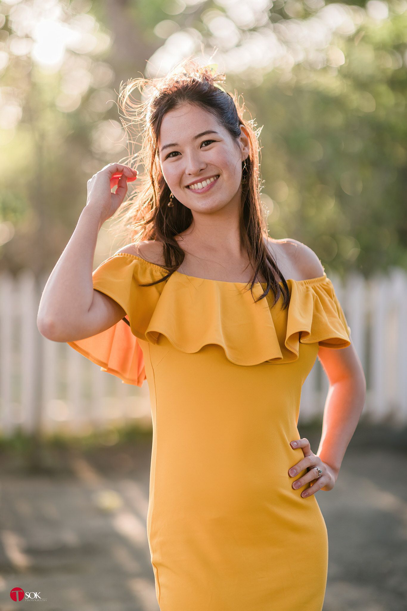 A woman in a yellow off-shoulder dress poses outdoors in golden sunlight.
