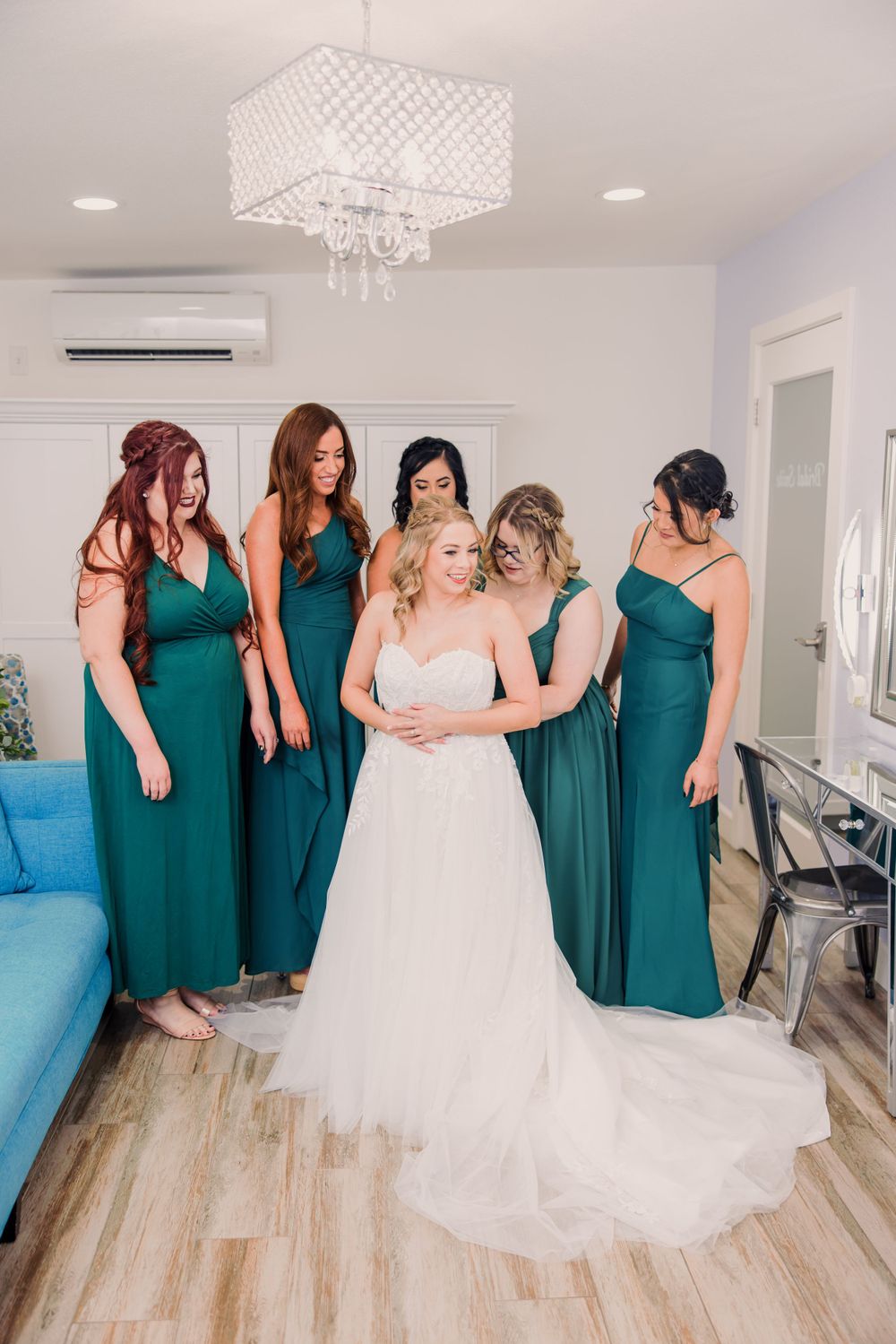 Bridesmaids in emerald green dresses help bride with wedding gown in bright indoor setting.