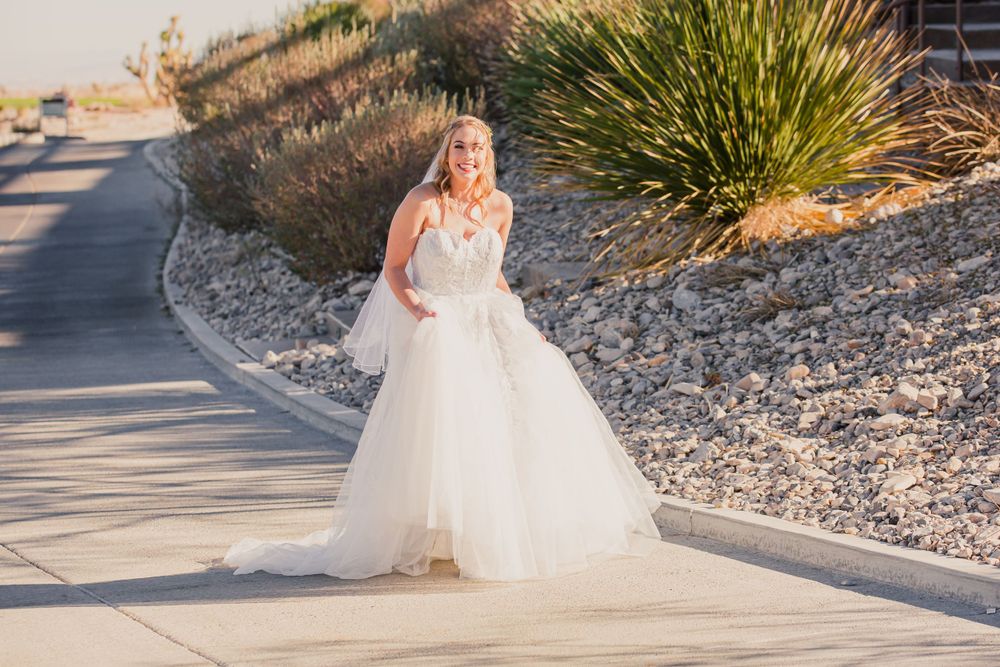 Bride in flowing white gown walks along desert garden pathway during sunset.