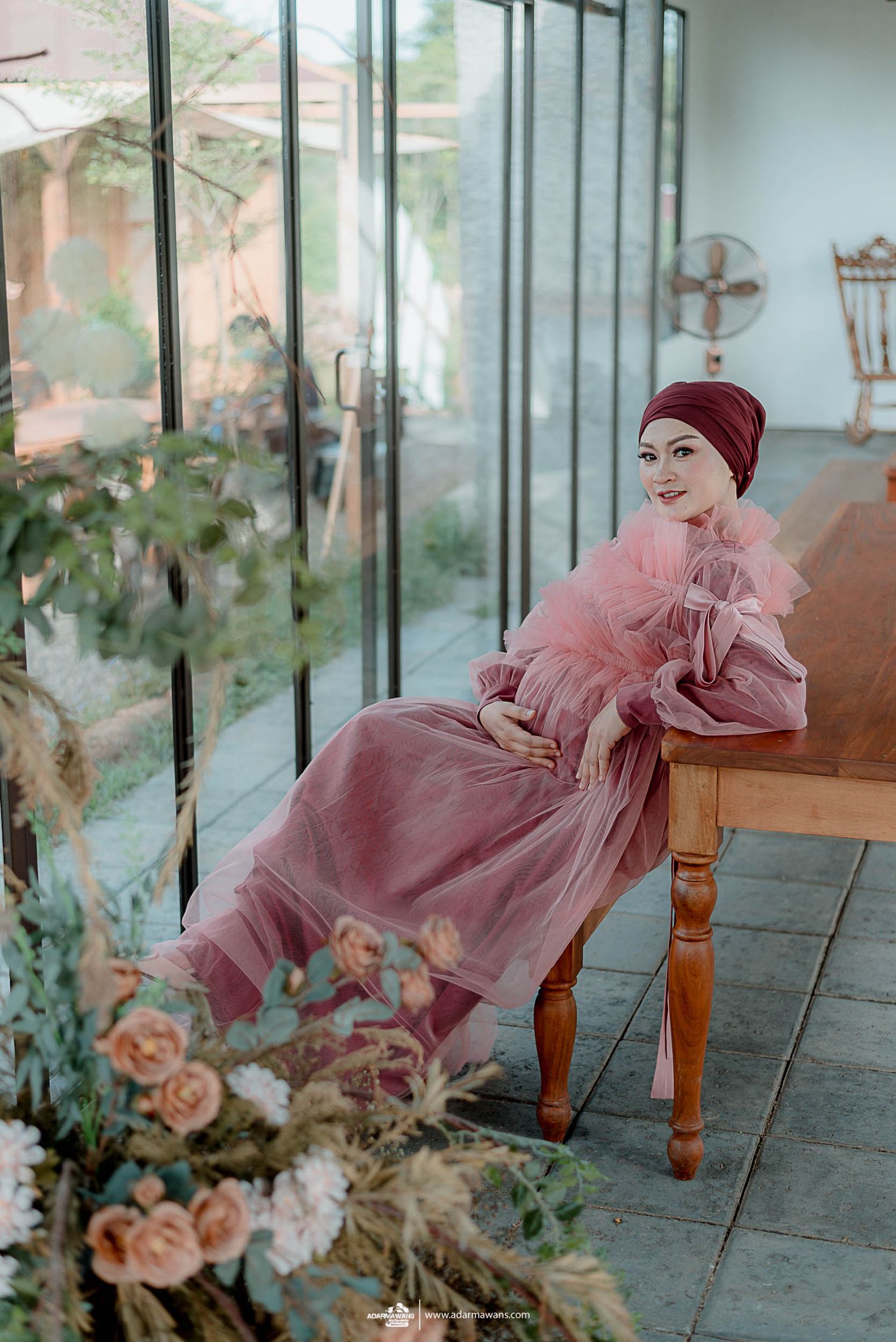 Person in flowing pink dress and red headpiece seated on wooden bench surrounded by flowers.