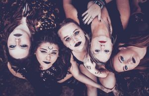 A group of people with gothic makeup lying in a circle formation with their heads together against a dark background.