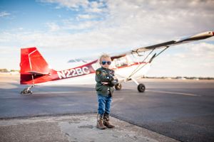 A small red and white aircraft parked on an airfield tarmac with child dressed as maverick in winter clothing standing beside it.