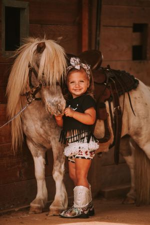 Young child in western attire poses with friendly pony inside rustic barn setting.