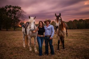 A couple stands with three majestic horses in a field at sunset with dramatic purple skies.