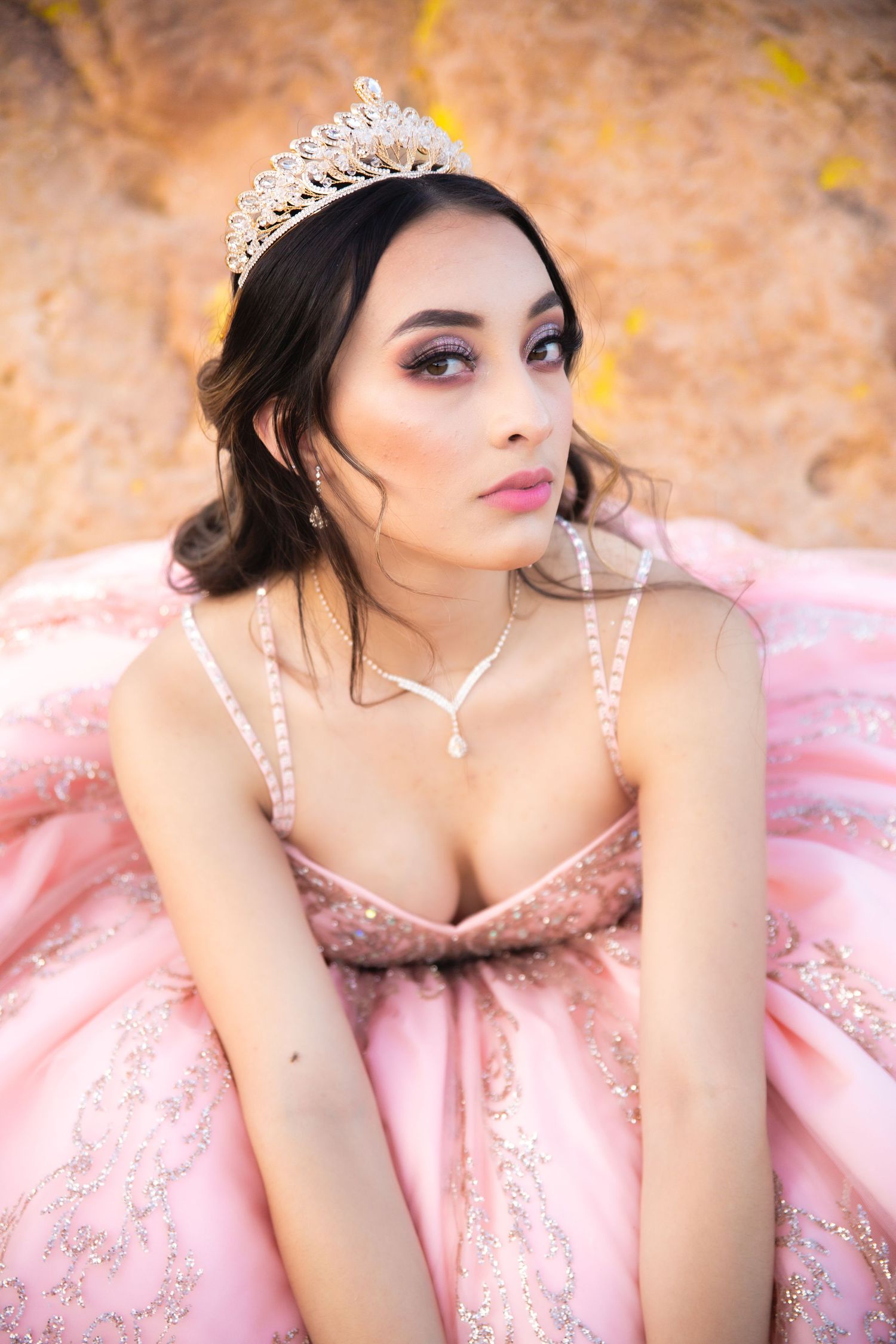 A woman in a sparkly pink ball gown and tiara poses outdoors against an autumn background.