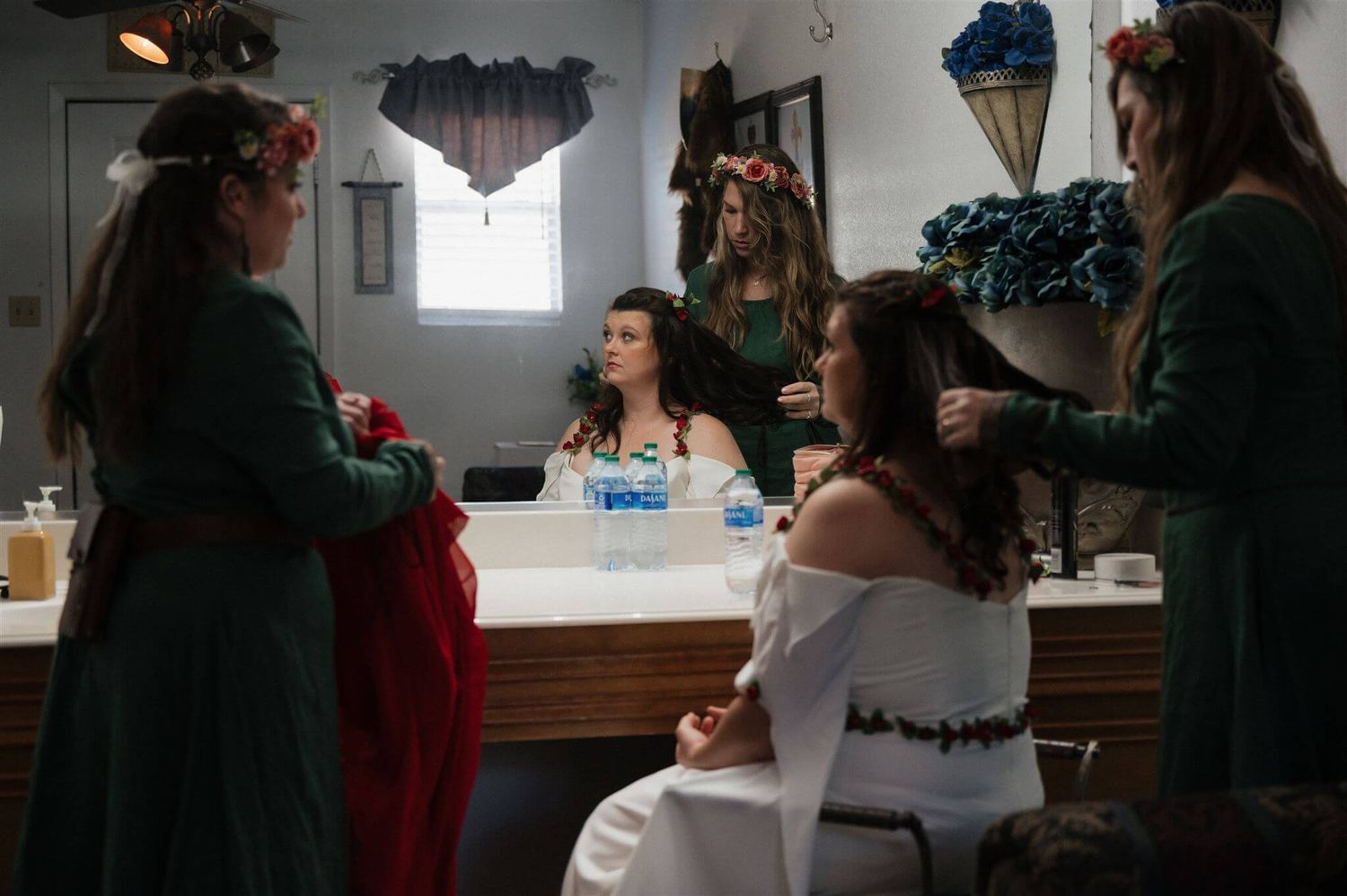 People in green medieval dresses gather around someone seated in white in a preparation room.