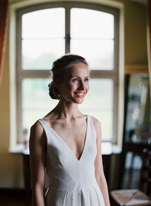 Bride wearing elegant V-neck wedding dress poses by window in natural light.
