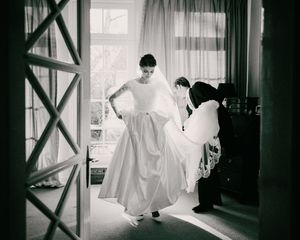 Two people adjusting a white wedding dress in a sunlit room.