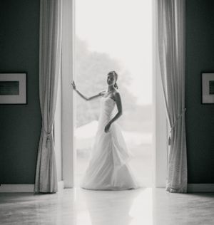 Silhouette of bride in flowing gown posed in bright window frame.