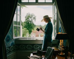 Person in hat standing by window with camera in dimly lit interior scene.
