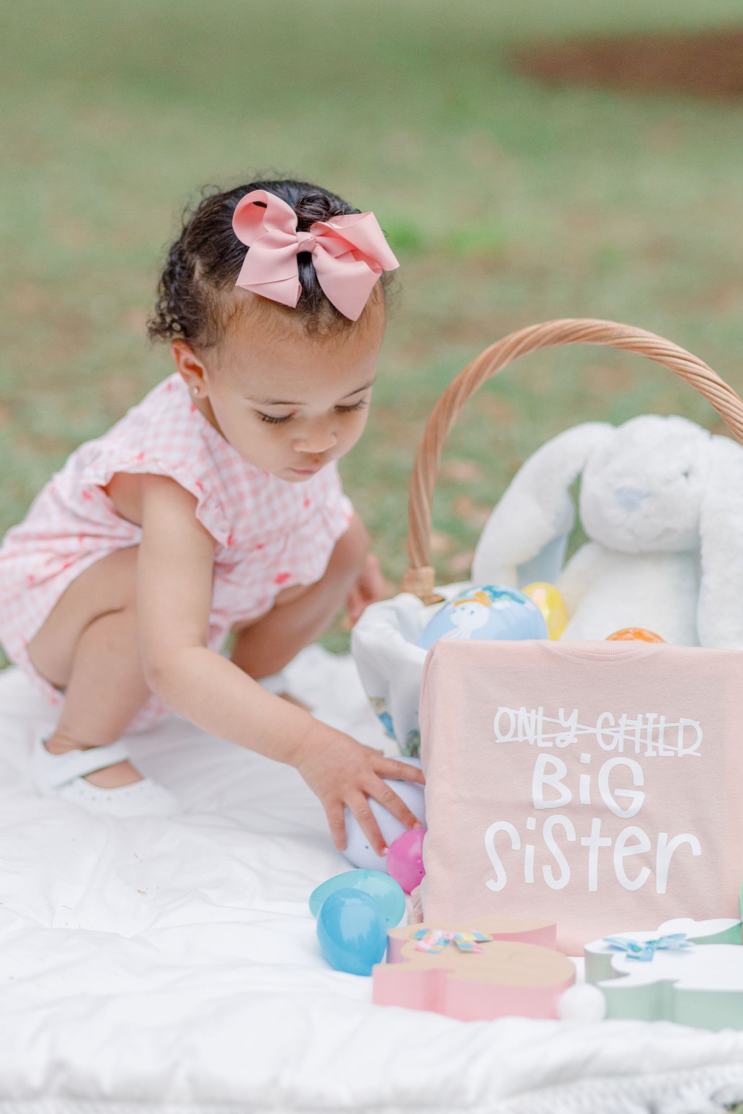 Baby girl in pink dress with bow playing with toys on white blanket next to 'Big Sister' sign.