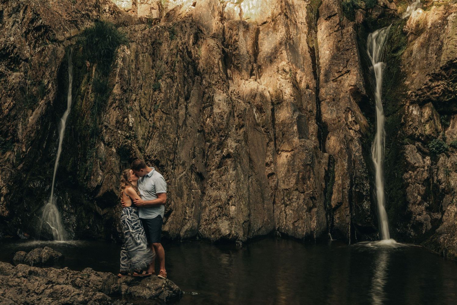 A couple stands by a scenic waterfall pool surrounded by rocky cliffs.