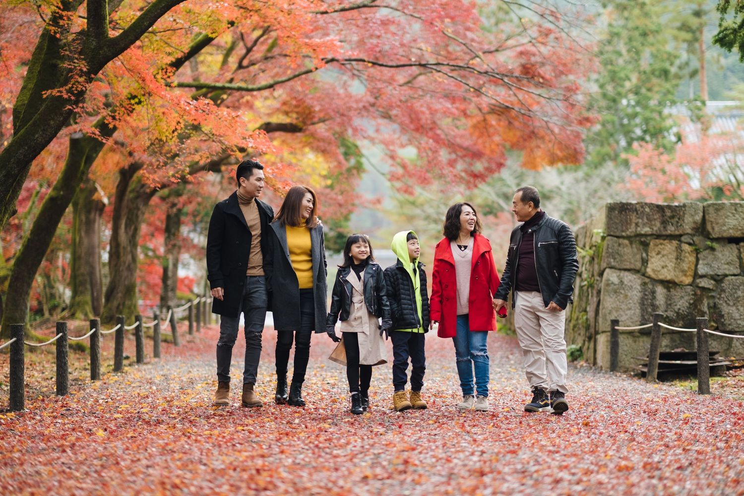 Family of six walking through a Japanese park path covered in red autumn leaves under colorful trees.