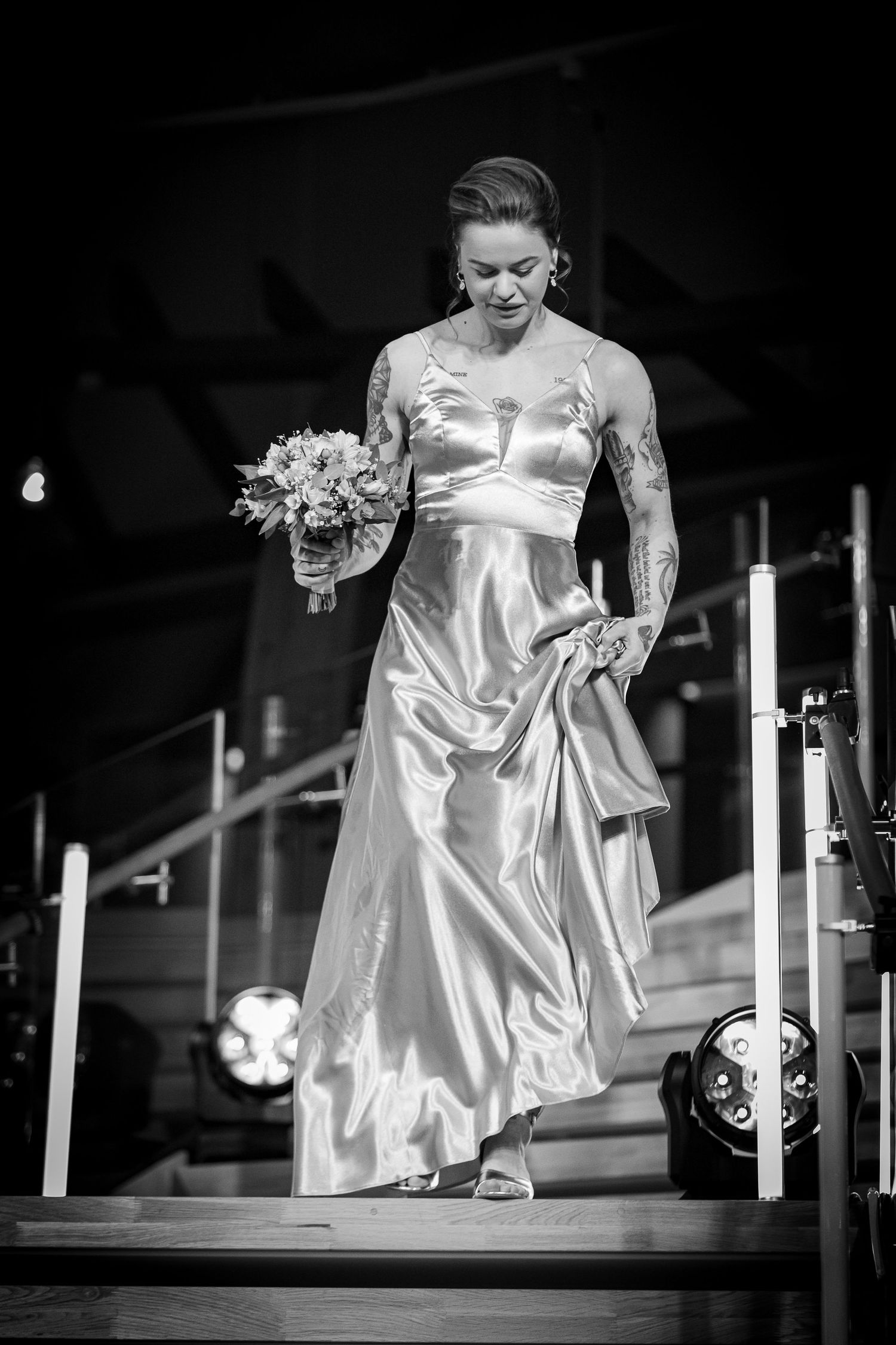 Black and white photo of tattooed bride in satin gown holding bouquet descending stairs.